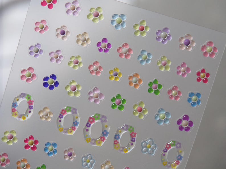 May include: A sheet of colorful flower stickers with a 3D effect. The stickers are arranged in rows and columns on a clear background. The flowers are in various colors, including pink, purple, blue, green, and yellow.