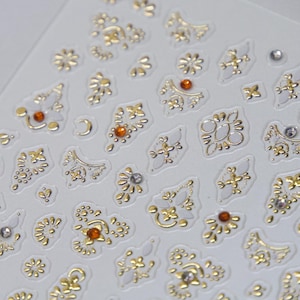 Puede incluir: Primer plano de una hoja de pegatinas decorativas con una variedad de dise&ntilde;os de filigrana dorados y plateados. Algunas pegatinas tienen peque&ntilde;os acentos de cristal naranja o transparente. Las pegatinas est&aacute;n sobre un fondo blanco.
