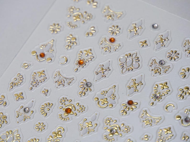 Puede incluir: Primer plano de una hoja de pegatinas decorativas con una variedad de dise&ntilde;os de filigrana dorados y plateados. Algunas pegatinas tienen peque&ntilde;os acentos de cristal naranja o transparente. Las pegatinas est&aacute;n sobre un fondo blanco.
