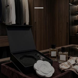 May include: A white ceramic flower-shaped stone placed in front of an open black gift box with foam inserts, accompanied by two glass perfume bottles and a patterned silk scarf. The text reads: SOPHISTICATION IN EVERY STONE and Artisanal ceramic stones designed for the elite professional's personal sanctuary.