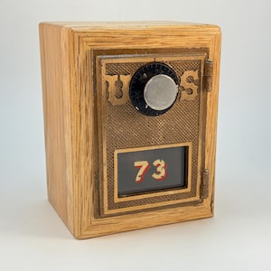 May include: A wooden box with a metal front panel resembling a safe. The front features a combination dial, the word "TUES" and the number "73". The box is made of light-colored wood with a natural grain.
