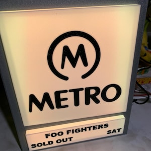 May include: A white sign with a black circle containing the letter "M" and the word "METRO" in black lettering. Below the word "METRO" is a smaller white sign with the text "FOO FIGHTERS SOLD OUT SAT" in black lettering.