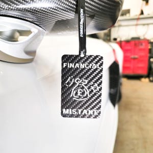 May include: A black and white carbon fiber tag with the words "FINANCIAL MISTAKE" and a graphic of a money bag with a pound symbol. The tag is hanging from a black strap that says "FORGEDMOTION".