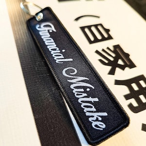 Peut inclure: Un porte-clés rectangulaire en tissu noir avec un anneau en métal argenté. Le porte-clés présente le texte brodé en blanc Financial Mistake dans une police cursive. Les bords sont finis avec une bordure cousue noire pour la durabilité.
