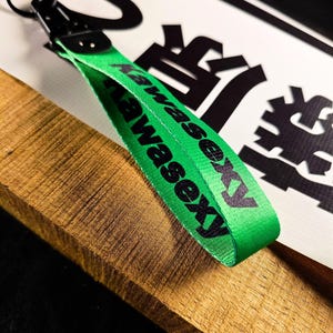 May include: A green lanyard with the text "Kawasexy" in black. The lanyard has a black metal clasp and key ring. The background includes a wooden surface and a white sign with black Japanese characters.