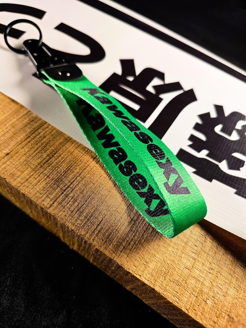 May include: A green lanyard with the text "Kawasexy" in black. The lanyard has a black metal clasp and key ring. The background includes a wooden surface and a white sign with black Japanese characters.