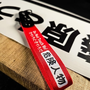 Puede incluir: Un llavero de tela roja con el texto "Do Not Touch Me!" y caracteres japoneses. El llavero tiene un cierre de metal negro y una franja blanca con texto japonés. El fondo incluye caracteres japoneses y una superficie de madera.
