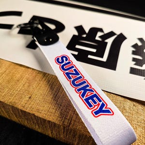 Op de afbeelding: Witte stoffen sleutelhanger met het woord "SUZUKEY" in blauwe en rode letters. De sleutelhanger heeft een zwarte metalen sluiting. De achtergrond bevat Japanse karakters en een houten oppervlak.