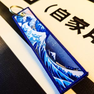 May include: A rectangular fabric keyring featuring a vibrant blue and white graphic print of a large ocean wave and a mountain peak. The keyring has a dark blue stitched border and a silver metal ring attached through a reinforced eyelet at the top.