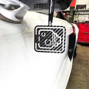 May include: A black and white carbon fiber car air freshener with a white outline of a camera. The air freshener is hanging from a black lanyard with the text "FORGEDNOTION" printed on it.