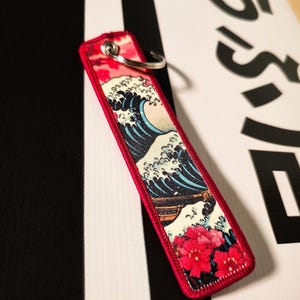 Può includere: Un portachiavi rettangolare rosso con un design a onda giapponese e fiori rossi. Il portachiavi ha un portachiavi argentato e un bordo rosso. Il design utilizza sfumature di blu, bianco e rosso.