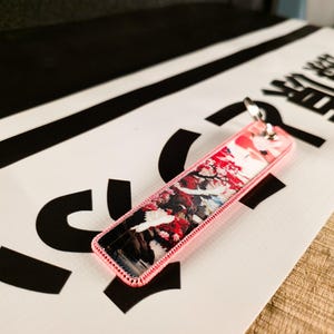 May include: A rectangular keychain with a floral design and a pink border. The keychain has a silver key ring attached. The background is a white surface with black shapes and Japanese characters.