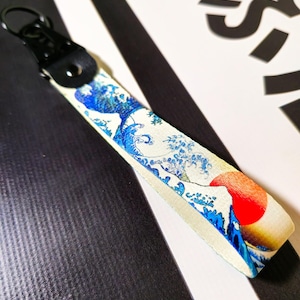 Puede incluir: Un llavero blanco y azul con una imagen impresa de una ola y un sol rojo. El llavero tiene una correa de cuero negro y un anillo de metal.