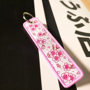 May include: Embroidered keychain with a pink floral pattern on a white background, framed by a pink border. The keychain has a silver metal ring for attachment. The design features cherry blossom flowers.