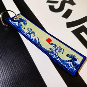 Puede incluir: Un llavero bordado en azul y blanco con una representación de la Gran Ola de Kanagawa, una famosa estampa japonesa en madera de Hokusai. El llavero tiene un círculo rojo que representa el sol naciente.