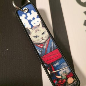 Puede incluir: Un llavero rectangular con un gato con kimono. El gato tiene pelaje blanco y ojos azules, vistiendo un kimono azul y rojo. La parte superior del llavero tiene un patrón azul y blanco, y la inferior un bodegón de comida.