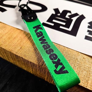 May include: A green keychain strap with the word "Kawasexy" printed in black. The strap is attached to a black metal clasp and ring. The background includes a white sign with black Japanese characters.