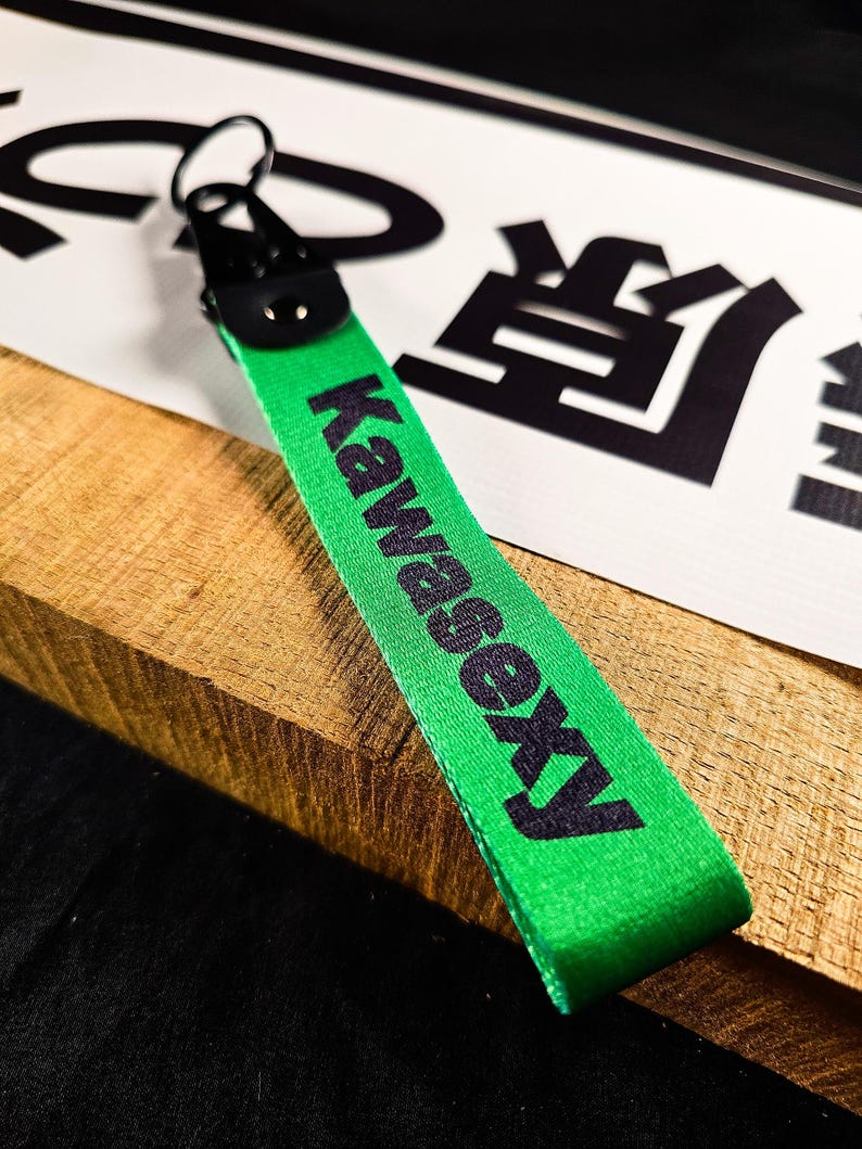 May include: A green keychain strap with the word "Kawasexy" printed in black. The strap is attached to a black metal clasp and ring. The background includes a white sign with black Japanese characters.