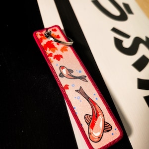 Puede incluir: Un llavero rectangular con borde rojo y fondo rosa. El llavero presenta dos ilustraciones de peces koi, hojas de arce rojas y burbujas azules. Un llavero plateado está adjunto en la parte superior.