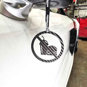 May include: A carbon fiber car accessory with a circular design featuring a crossed-out soda cup and burger. The accessory hangs from a black strap with the text "FORCEDMOTION". The accessory is hanging on a white car.