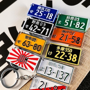 Peut inclure: Collection de porte-clés plaques d'immatriculation japonaises de différentes couleurs, dont le bleu, le vert, l'orange et le jaune. Chaque porte-clés présente des combinaisons de chiffres uniques et des caractères japonais. Les porte-clés sont attachés à des anneaux argentés.