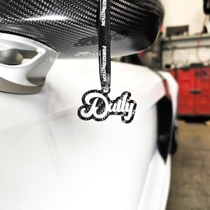 May include: A black and white carbon fiber car charm with the word "Daily" in a stylized font. The charm is hanging from a black ribbon with the words "Forged Motion" printed on it.