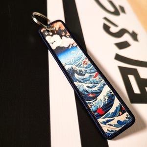 Puede incluir: Llavero bordado con diseño de olas japonesas, borde azul marino y anilla plateada. La obra de arte representa una montaña, nubes y olas rompiendo en tonos azules, rojos y blancos.