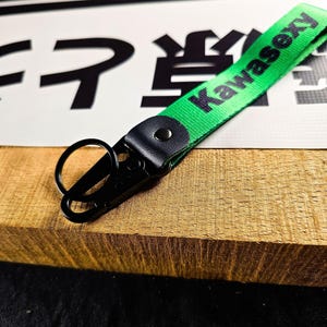 May include: A green fabric keychain with the word "Kawasexy" printed in black. The keychain has a black metal clasp and a black leather accent. The background includes Japanese characters and a wooden surface.