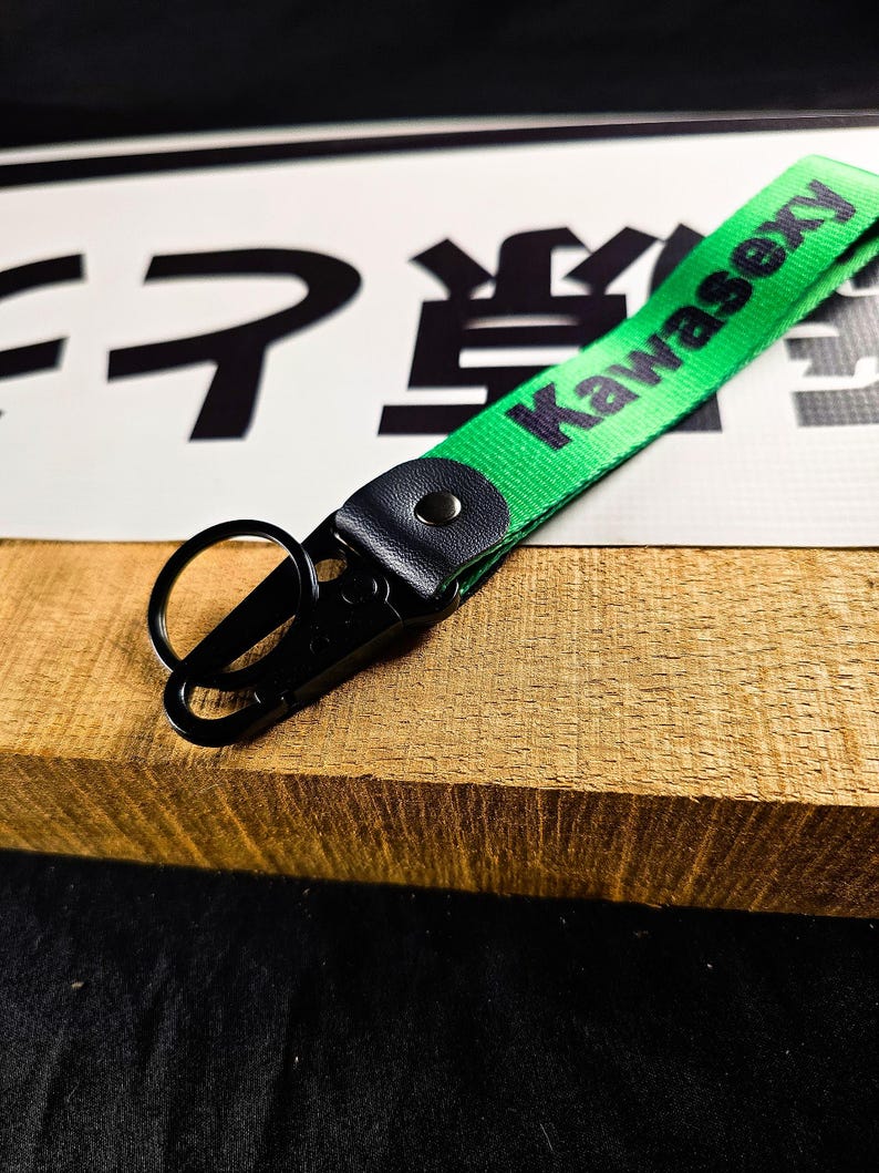 May include: A green fabric keychain with the word "Kawasexy" printed in black. The keychain has a black metal clasp and a black leather accent. The background includes Japanese characters and a wooden surface.