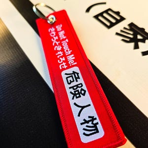 Könnte beinhalten: Ein rechteckiger roter Stoffschlüsselanhänger mit einem Metallring. Der Schlüsselanhänger zeigt weißen Text "Do Not Touch Me!" und japanische Schriftzeichen sowie einen weißen Aufnäher mit dem japanischen Text "危険人物".