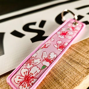 Puede incluir: Llavero floral rosa bordado con diseño de flores de cerezo. El llavero rectangular tiene centros rojos, pétalos blancos y un borde rosa. Tiene un llavero plateado y está sobre una superficie de madera.