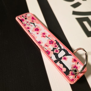 Pode incluir: Um porta-chaves retangular com uma borda rosa e um padrão floral de flores rosa sobre um fundo branco. O porta-chaves tem um anel de metal prateado. O fundo é preto e branco.