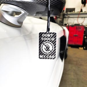 May include: A black and white car air freshener with a carbon fiber pattern. The air freshener has a white hand with a stop sign around it and the text "Don't Touch My Car".