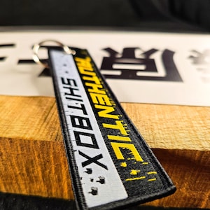May include: Black and white rectangular keychain tag with the words "AUTHENTIC SHITBOX" in yellow and white lettering. The tag is attached to a silver metal keyring. The background is a wooden surface.