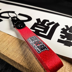 Puede incluir: Llavero de tela roja con cierre metálico negro. El llavero tiene el texto "OSAKA LOOP CRAZY GANG" en blanco y negro. El fondo incluye caracteres japoneses y una superficie de madera.