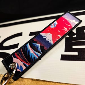 May include: A rectangular black keychain tag featuring a vibrant Japanese landscape design. The artwork depicts a red sun, white bird, and blue mountains. A silver key ring is attached to the tag.