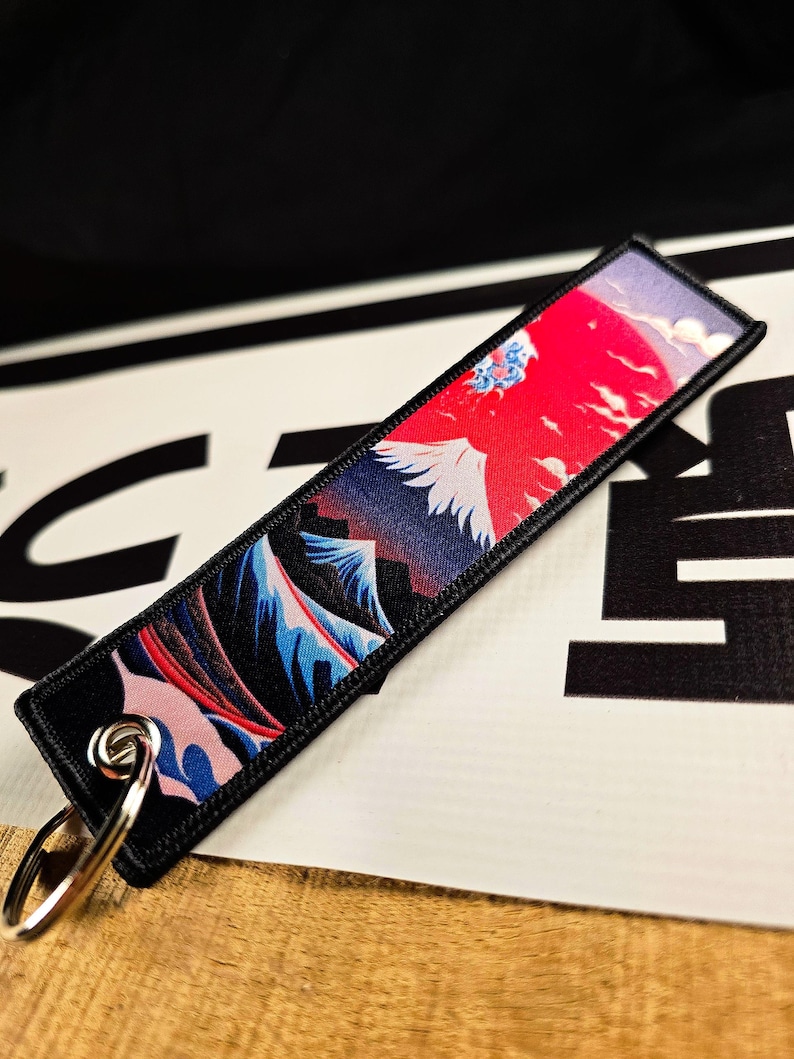 May include: A rectangular black keychain tag featuring a vibrant Japanese landscape design. The artwork depicts a red sun, white bird, and blue mountains. A silver key ring is attached to the tag.
