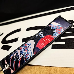May include: A rectangular keychain tag featuring a Japanese wave design with a red sun, blue waves, and red accents. The tag has a black border and a silver metal ring for attachment.