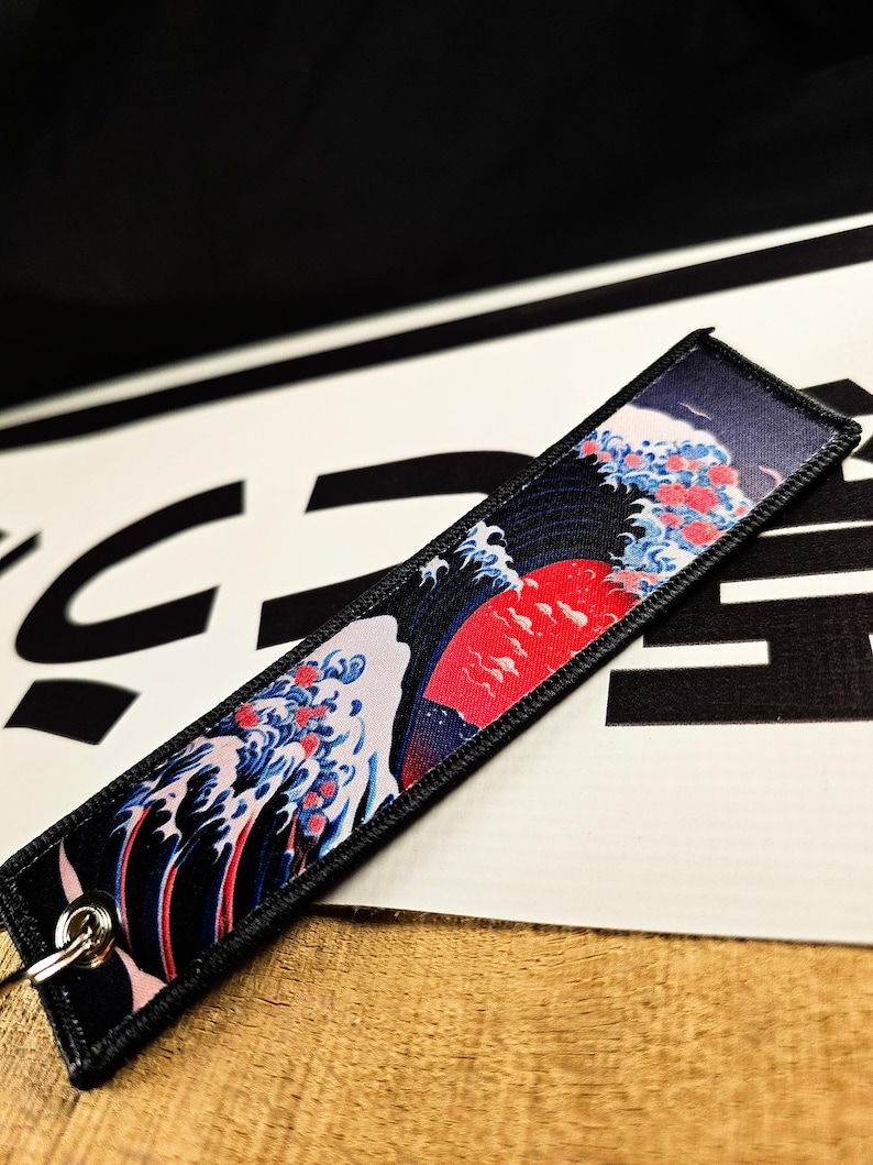 May include: A rectangular keychain tag featuring a Japanese wave design with a red sun, blue waves, and red accents. The tag has a black border and a silver metal ring for attachment.