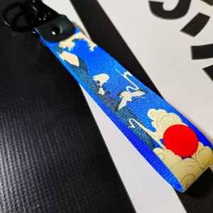 Puede incluir: Llavero azul y blanco con un círculo rojo y un diseño de montaña y ola de estilo japonés.
