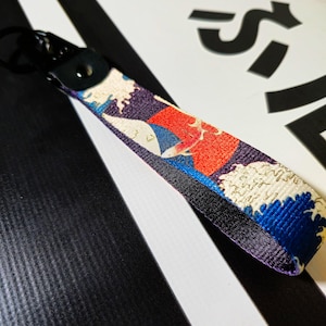 Puede incluir: Un llavero negro y azul con un diseño de ola japonesa colorido. El llavero tiene un anillo de metal y una correa negra.