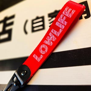 Może przedstawiać: Jasnoczerwony pasek do kluczy z tkaniny z białym napisem LOWLIFE wydrukowanym przetartą czcionką. Pasek jest przymocowany do czarnego metalowego karabińczyka z czarnym, teksturowanym łącznikiem z imitacji skóry zabezpieczonym metalowym nitem.