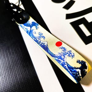 Puede incluir: Un llavero blanco y azul con un diseño de ola japonesa y un círculo rojo. El llavero tiene un anillo de metal negro.