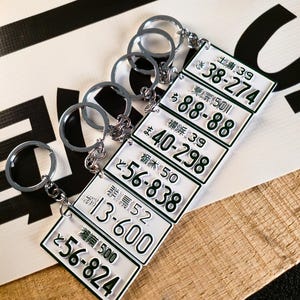 Puede incluir: Una colección de llaveros con miniaturas de matrículas japonesas. Cada llavero presenta una placa blanca con texto negro y un anillo plateado. Las placas muestran caracteres japoneses y combinaciones de números.