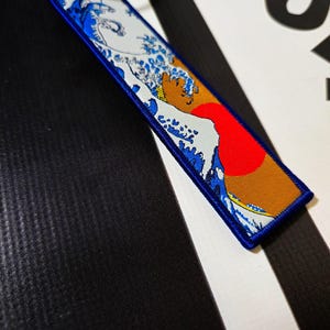 Puede incluir: Un llavero de tela azul y blanco con una representación de la Gran Ola de Kanagawa, una famosa estampa japonesa en madera de Hokusai. La ola está representada en azul y blanco, con un sol rojo saliendo en el fondo. El llavero tiene un borde azul y un anillo plateado.