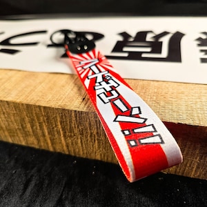 May include: A red and white wrist strap with Japanese text and a rising sun design. The strap has a black clasp and is resting on a wooden surface.