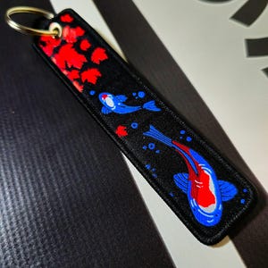 May include: Black key chain featuring a red and blue koi fish design. The fish are swimming through a field of red leaves.