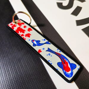 Puede incluir: Un llavero blanco con un diseño de pez koi rojo y azul. El llavero tiene un anillo plateado y hojas de arce rojas en el fondo.
