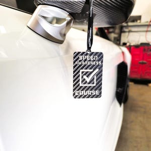 May include: A black and white car air freshener with a carbon fiber pattern. The air freshener has a checkmark and the text "SPEED AWARENESS COURSE".