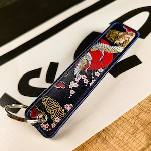 Pode incluir: Uma etiqueta de chaveiro azul marinho com um design de guindaste. A etiqueta apresenta um sol vermelho, flores de cerejeira brancas e detalhes dourados. Um porta-chaves prateado está preso.